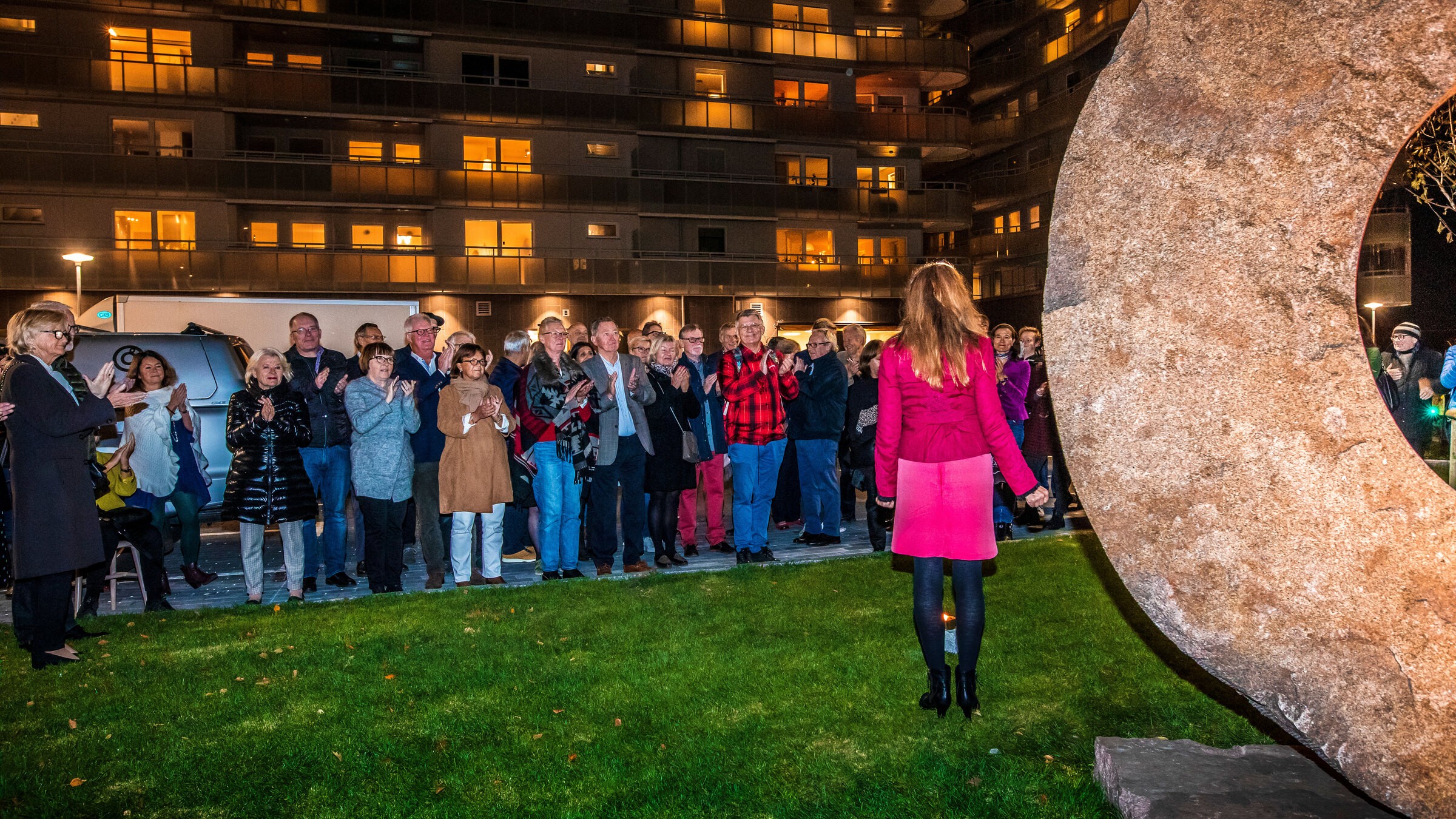 En kvinna i rosa klädsel talar till en folkmassa nära en stor stenstaty på natten.