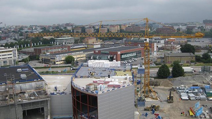 Byggarbetsplats med kranar, byggnader och material under en molnig himmel i ett stadsområde.