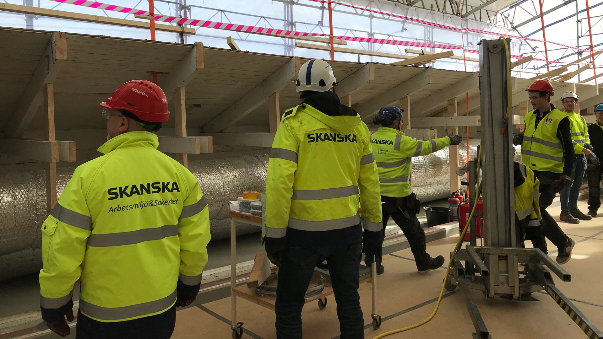 Byggnadsarbetare i högsynliga Skanska-jackor och hjälmar samarbetar på en byggarbetsplats.