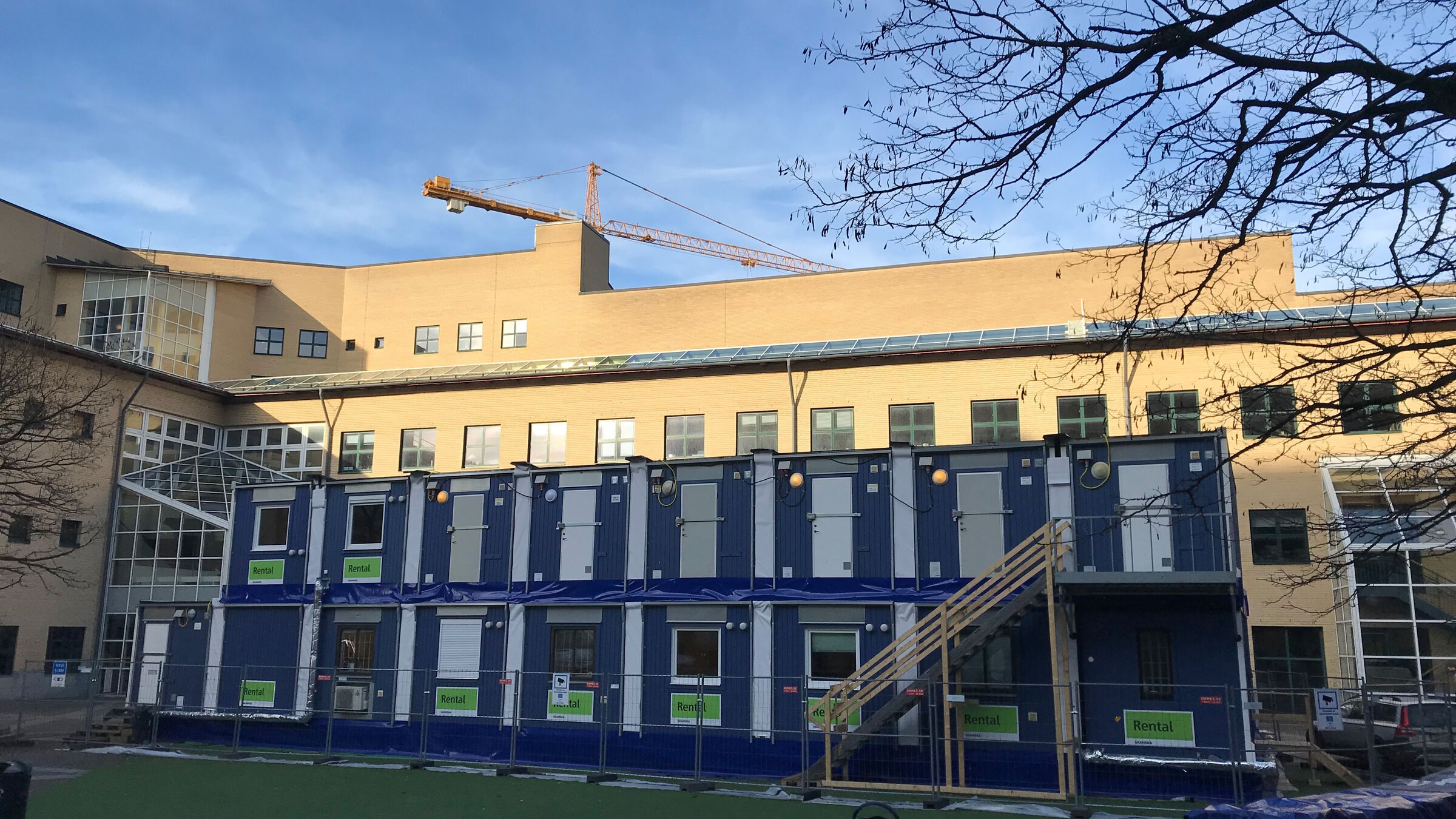 Byggarbetsplats med modulära enheter och kran nära en modern byggnad under en klar himmel.