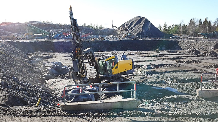 Tung maskin i arbete på byggarbetsplats Grävmaskin på ett stenbrottsområde, borrar i stenig terräng under starkt dagsljus, omgiven av jordhögar.