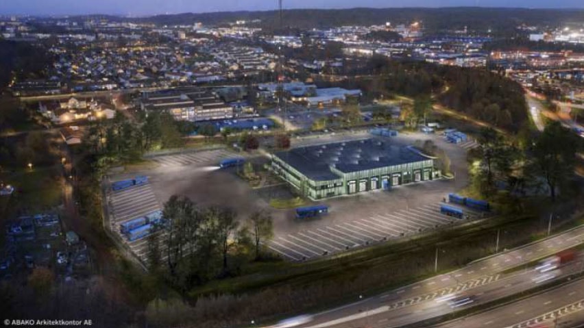 Vi bygger för en eldriven bussflotta och depån kommer ha plats för omkring 100 bussar. Flygvy av ett stort lager omgivet av parkeringsplatser och vägar i ett förortsområde vid skymning.