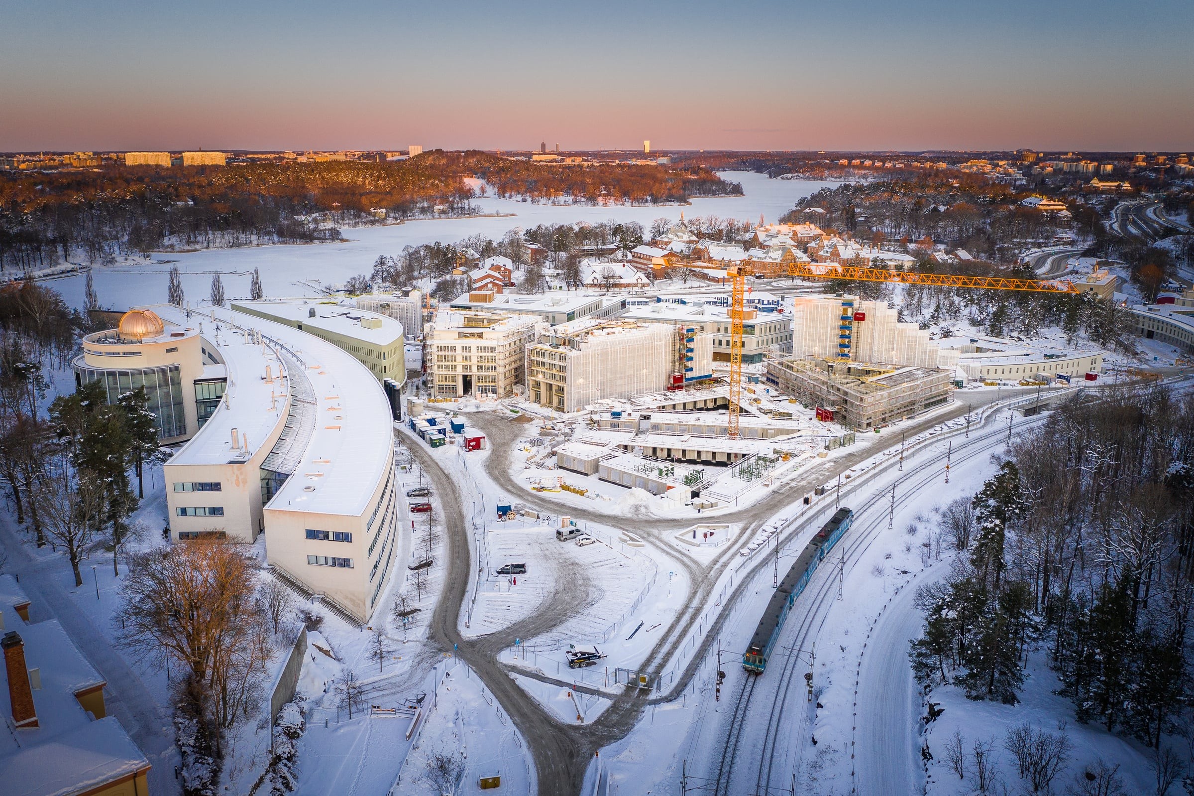 Flygvy av en snöig byggarbetsplats nära en frusen sjö, omgiven av vinterlandskap.