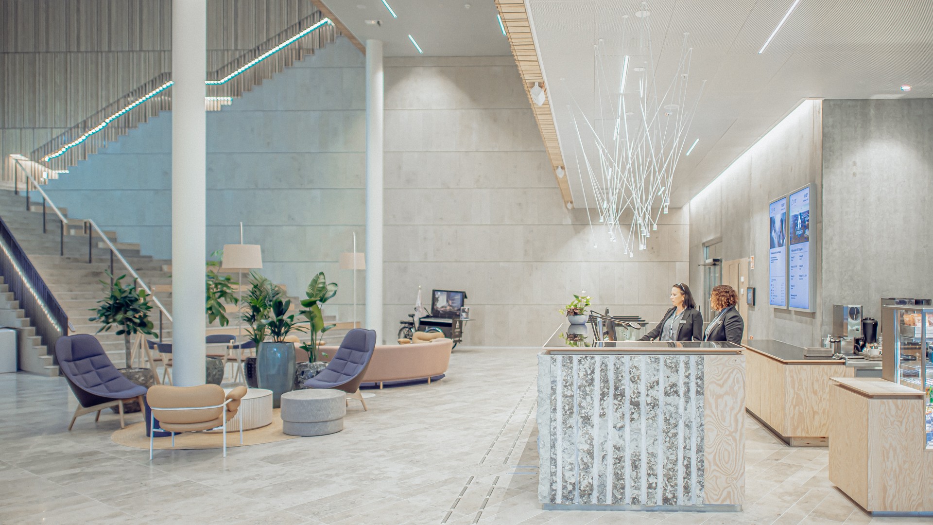 Till vänster skymtas den stora trappan, loungedel med fåtöljer och soffa. till höger syns receptionsdisken med två personer och en del av restaurangens diskar.