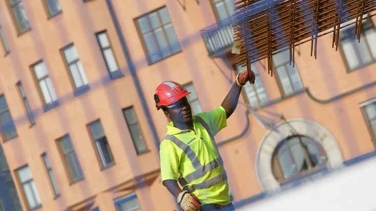 Clarion Sign Byggnadsarbetare i skyddsutrustning som leder en kran som lyfter armeringsjärn framför en byggnadsfasad.