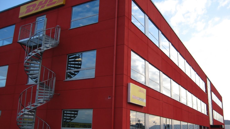 DHL Röd DHL-byggnad med spiraltrappa, stora fönster och en servicestationsskylt, under en blå himmel.