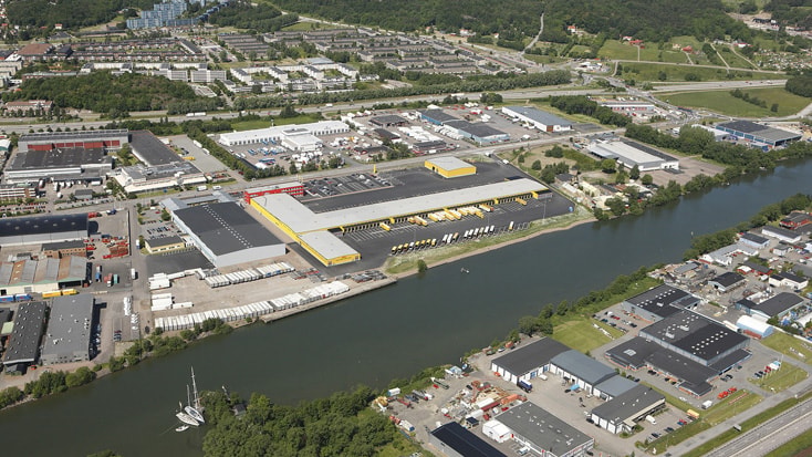 DHL Flygvy över ett industriområde med lagerbyggnader och en flod, omgivet av grönska och vägar.