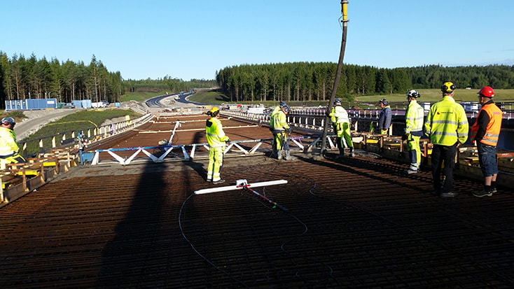 Byggnadsarbetare på en broplats övervakar betonggjutning, omgivna av skog och motorväg.