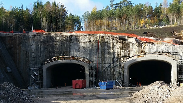 Byggarbetsplats med två stora tunnelentréer omgivna av byggnadsställningar och skogsområde.