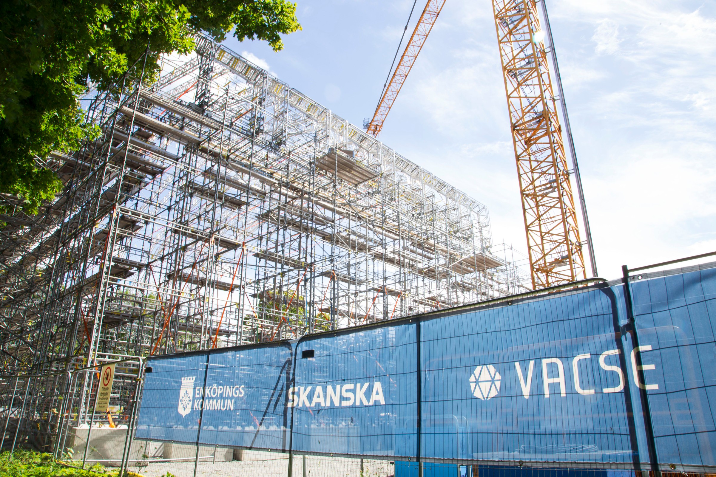 Ställning som bildar väggar och tak monteras med hjälp av em byggkran. I förgrunden syns stängsel med skyltning och loggorna Skanska och Vacse.