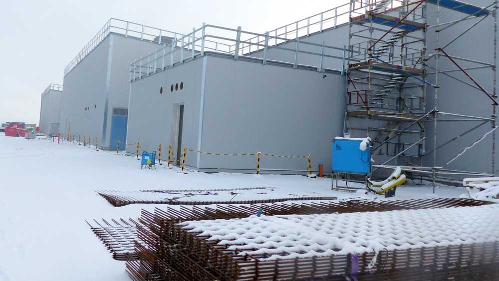 Snötäckt byggarbetsplats med byggnadsställningar, metallstänger och industribyggnader på vintern.