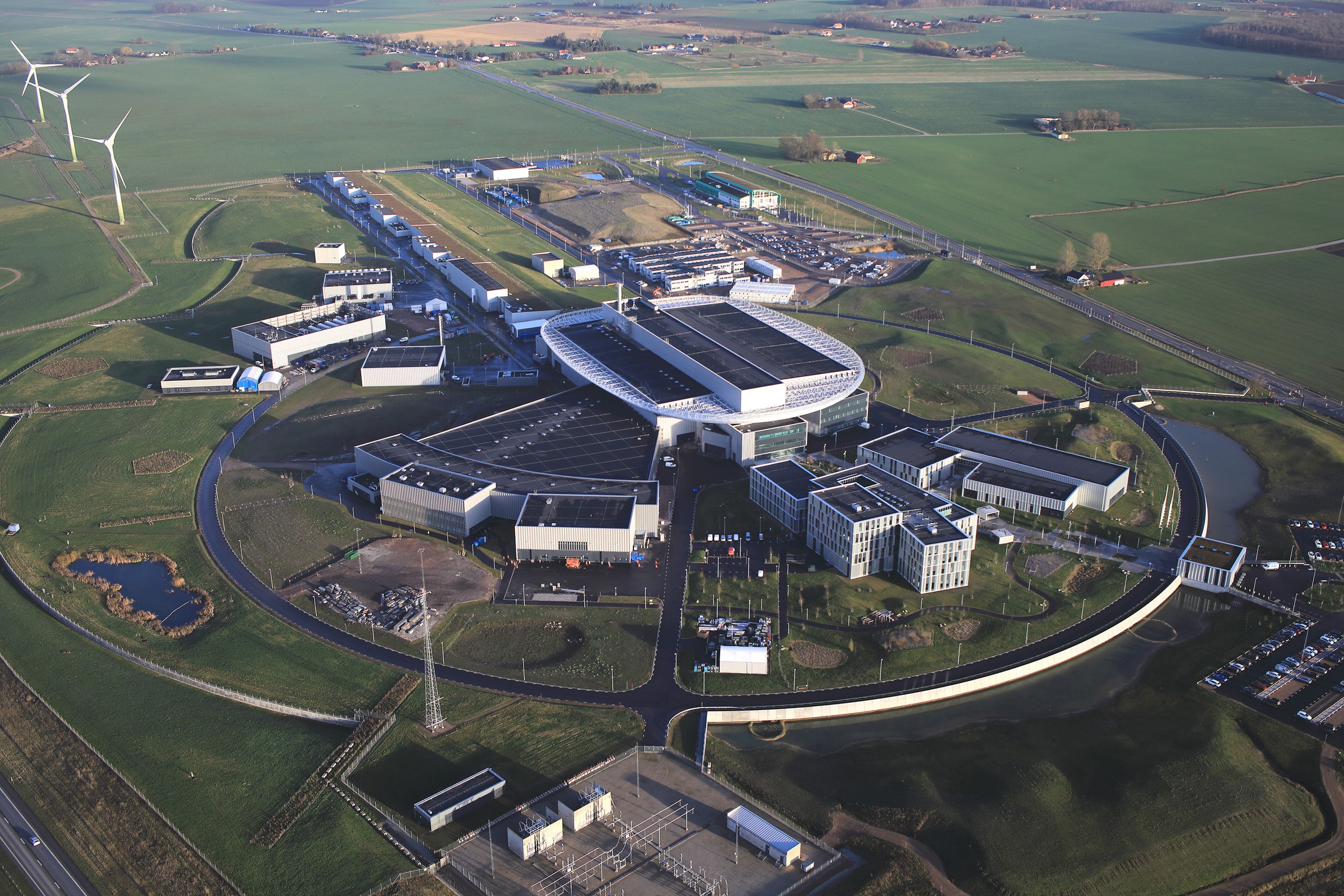 I december 2021 lämnar Skanska över helheten för byggnaderna till ESS. Foto: Perry Nordeng/ESS Flygvy över en modern forskningsanläggning omgiven av gröna fält och vindkraftverk.