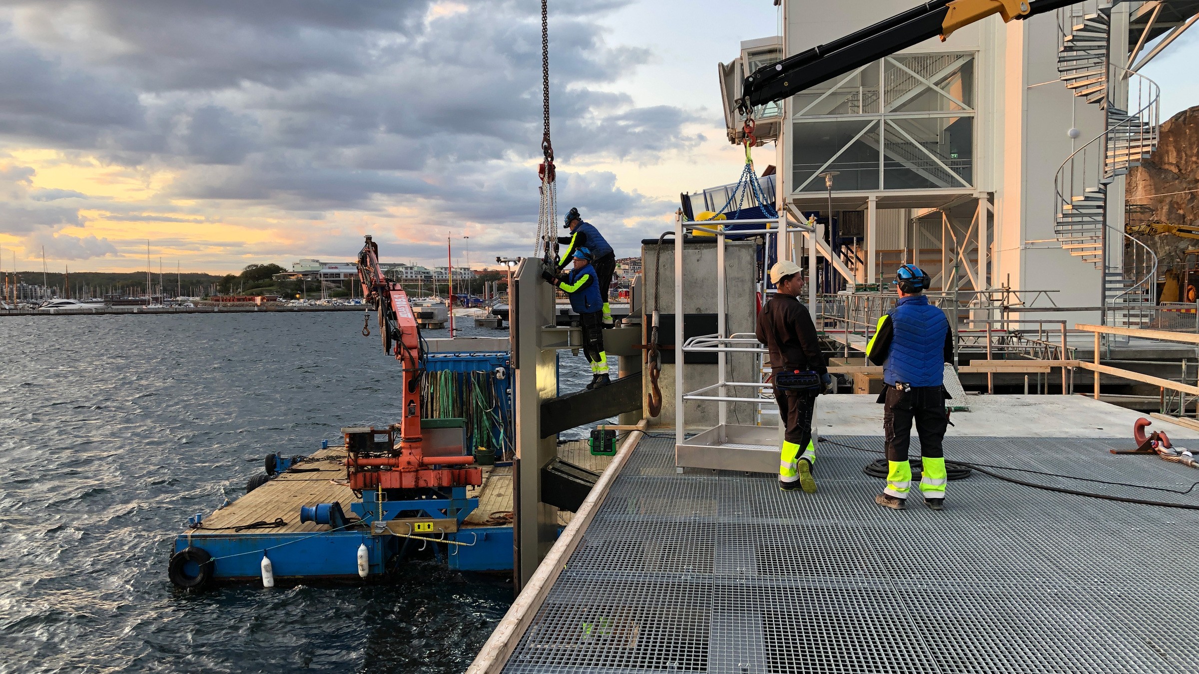 All pålning har skett från provisoriska pålbryggor från land. Arbetare installerar utrustning på en brygga vid havet, med hjälp av kranar och båtar under en molnig himmel.