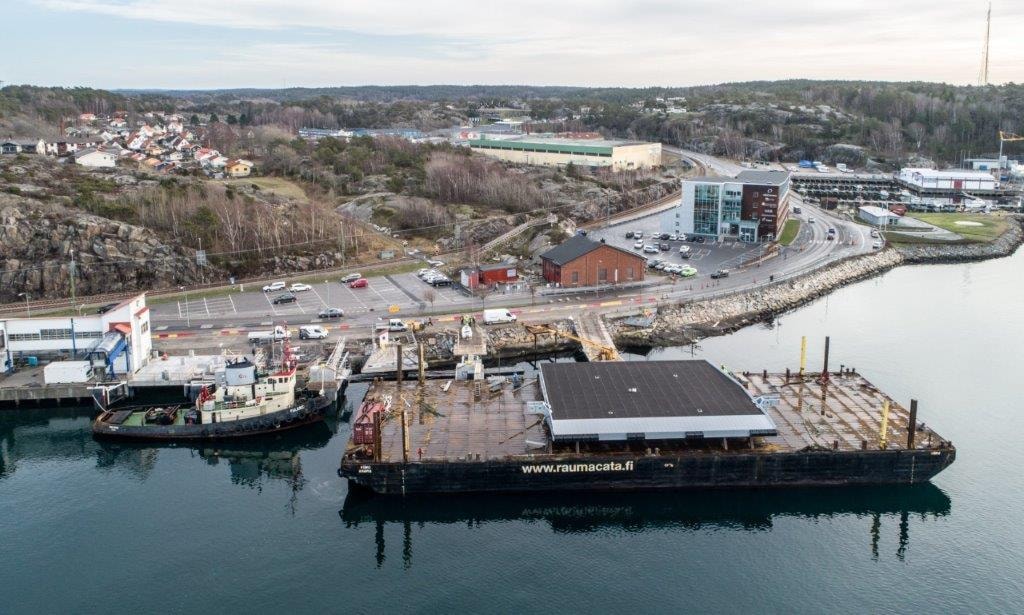 Den nya rampen anlände i januari till färjeläget. Flygvy över en kustbrygga med en stor pråm och omgivande byggnader, vägar och grönområden.