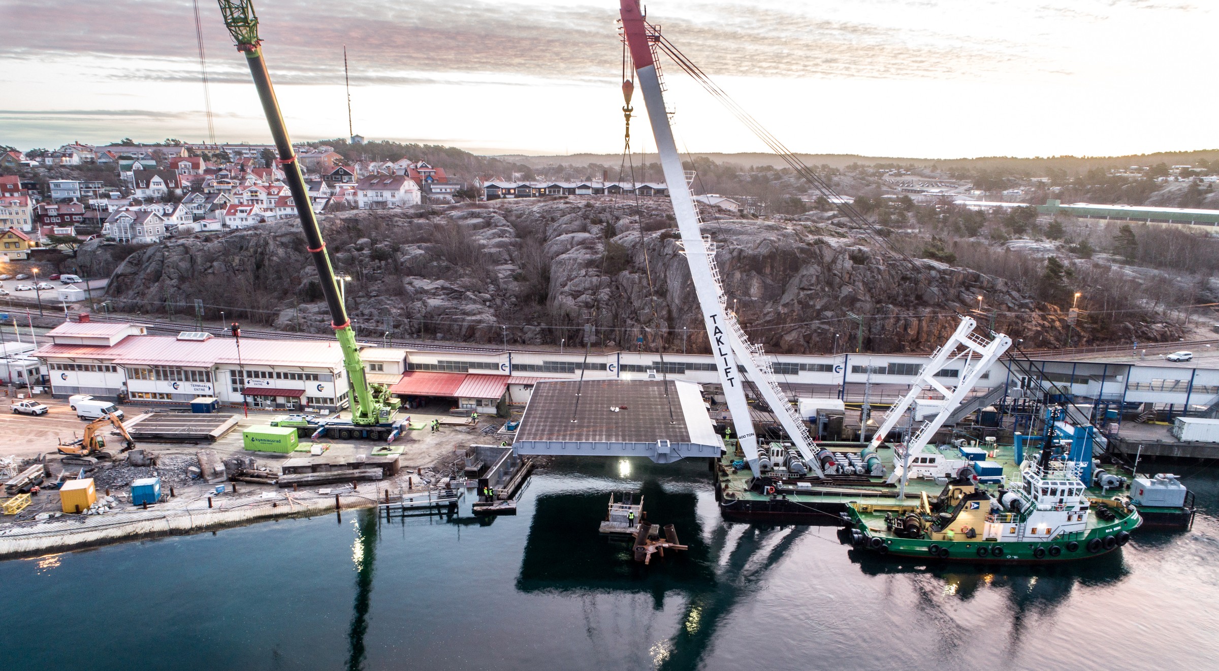 Färjeläget Torskholmen byggs om och får en ny ramp. Flygvy av kranar som lyfter en stor struktur vid vattenkanten, med ett stenigt landskap i bakgrunden.