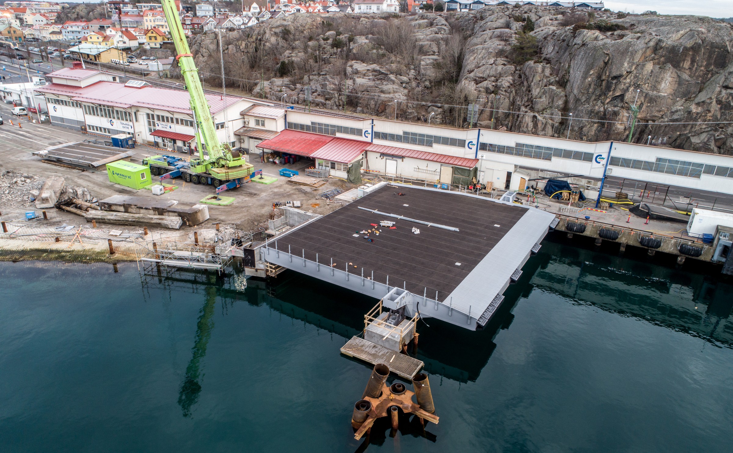 Med den nya rampen på plats är färjeläget nu redo att ta emot det nya laddhybridfartyget till sommaren 2019. Flygvy över en stor bryggplattform under konstruktion nära en stenig kustlinje och industriområde.
