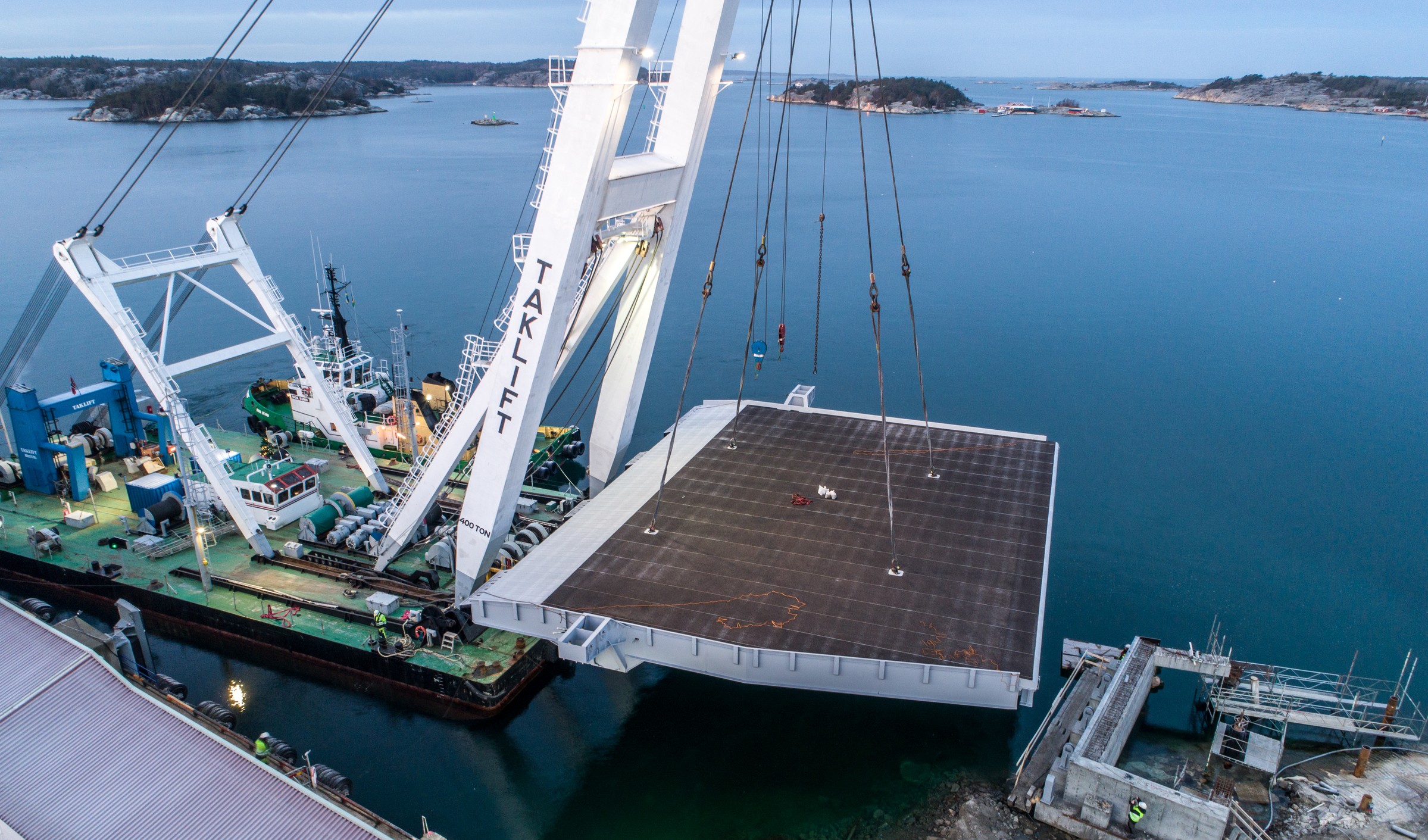 Monteringen skedde under ett två veckor långt båtstopp. En stor kran lyfter en massiv stålplattform över vatten, med en pråm och avlägsna öar synliga.