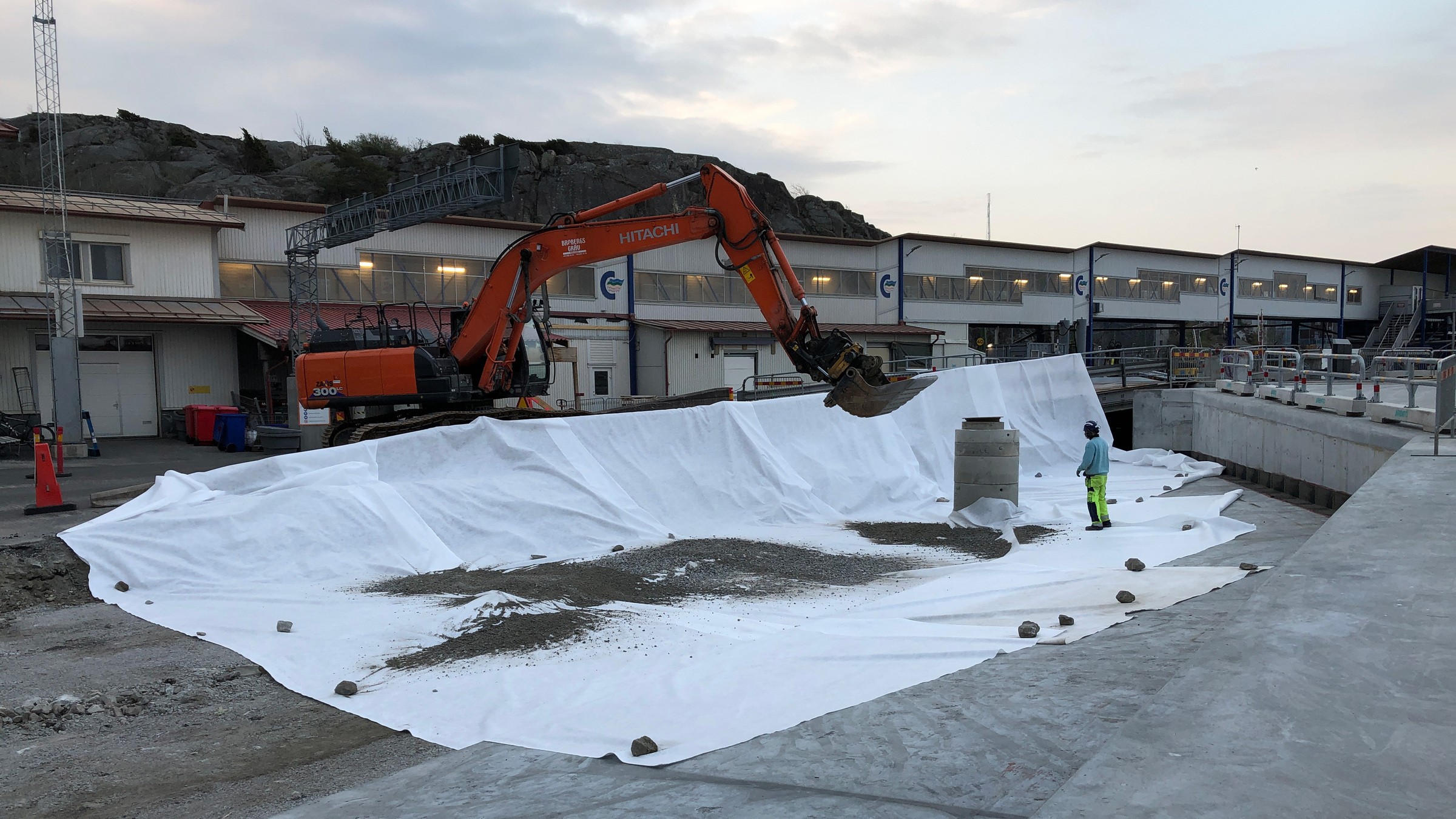 Projektet pågår till sommaren 2020. Grävmaskin lägger grus på en stor vit presenning vid en byggarbetsplats nära industribyggnader.