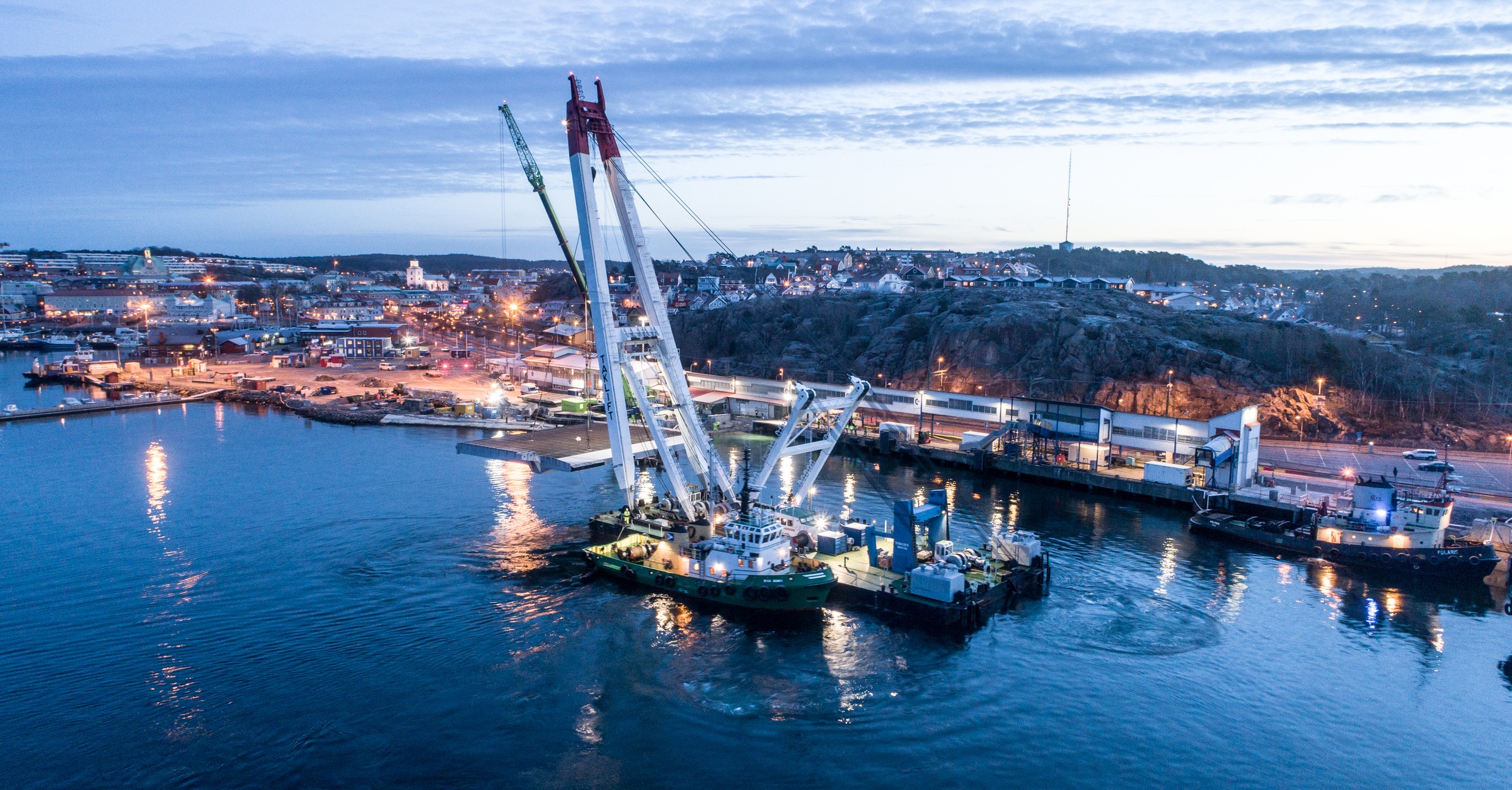 Rampen monterades på plats med hjälp av en norsk inhyrd kran. Flygvy av en stor kranpråm i en hamn vid skymning med stadsljus och klippig kustlinje i bakgrunden.