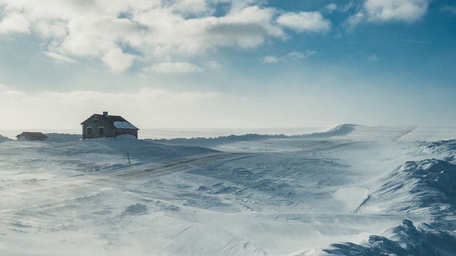 Ett ensamt hus ligger i ett vidsträckt, snötäckt landskap under en blå himmel med spridda moln.