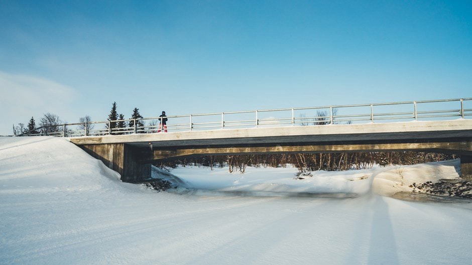 En snötäckt bro sträcker sig över ett fruset landskap under en klar blå himmel.