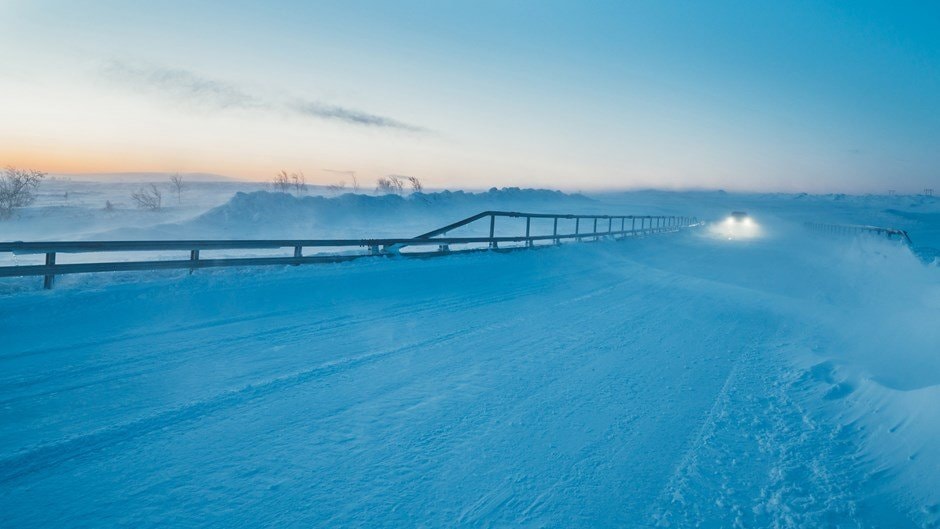 En snötäckt väg med yrande snö och strålkastare i fjärran under en vinter solnedgång.