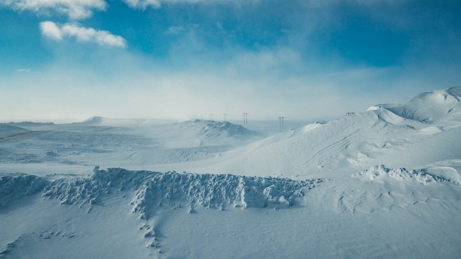 Snötäckt landskap med böljande kullar under en klarblå himmel; kraftledningar i fjärran.