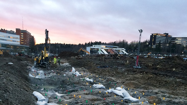 Byggarbetsplats i gryningen med maskiner, sandsäckar och byggnader i bakgrunden under en molnig himmel.