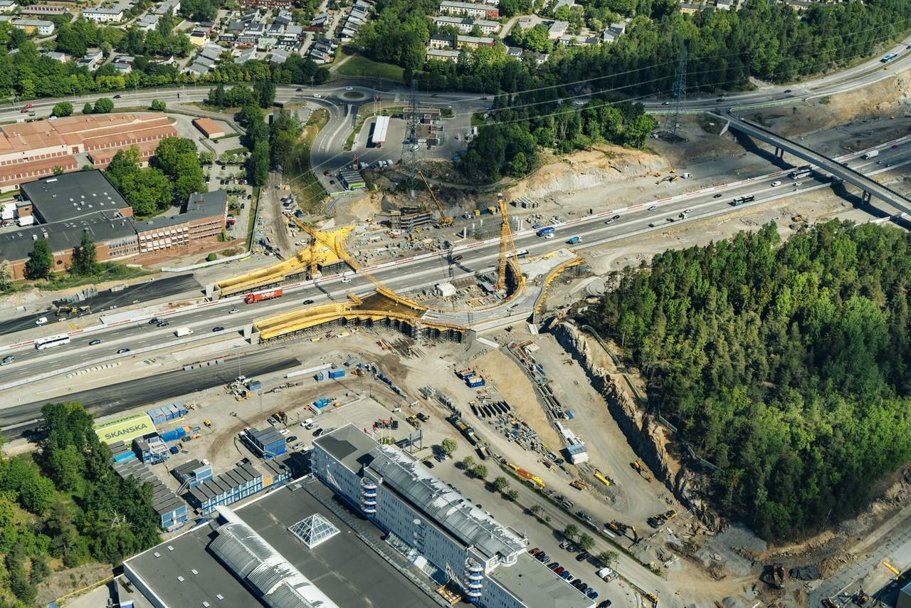 Flygvy över en vägbyggnadsplats med kranar, fordon och omgivande grönska.