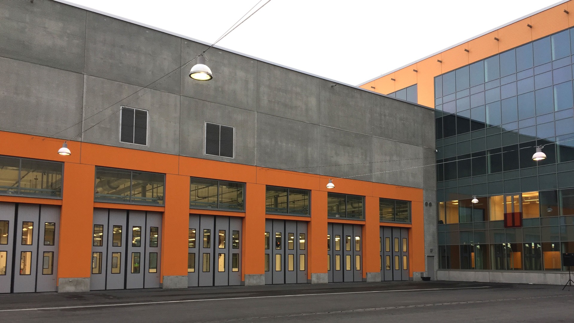 Från innergården leder portar in till depåns verkstad. Modern industribyggnad med ljusa orange accenter och stora garageportar, under klar himmel.