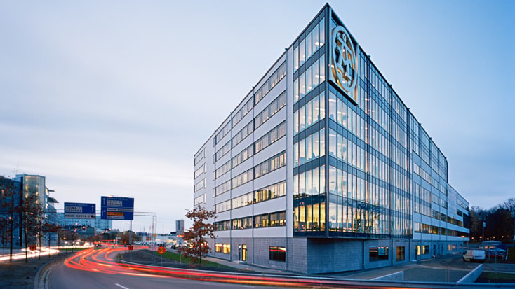 Modern office building with glass facade and prominent logo, situated along a busy street at dusk.