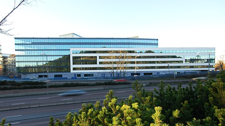 Modern glas-kontorsbyggnad med reflekterande fönster intill en trafikerad väg omgiven av grönska.