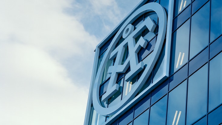 Modern glasfasad med en stor 3D abstrakt logotypskylt mot en blå himmel.