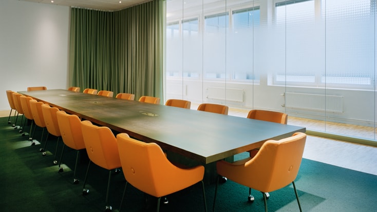Modern konferensrum med orange stolar och ett långt bord vid glasväggar och gardiner.