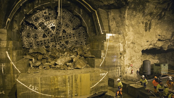 Hallandsås Tunnelborrmaskin i underjordisk byggarbetsplats med arbetare och bråte, svagt upplyst miljö.