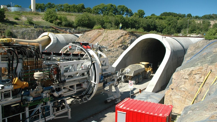 Hallandsås Tunnelborrmaskin och byggutrustning vid en tunnelingångsområde omgivet av träd.