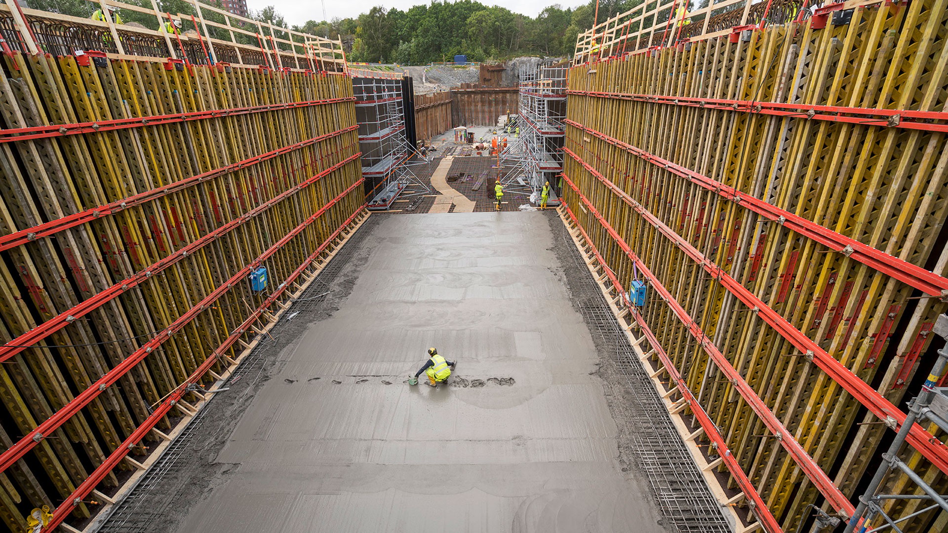 Betongen beräknas minska koldioxidutsläppen med minst 40 procent. Betongbil och kranar parkerade vid arbetsplatsen. Byggnadsarbetare jämnar ut betong mellan stora formpaneler på en byggarbetsplats.