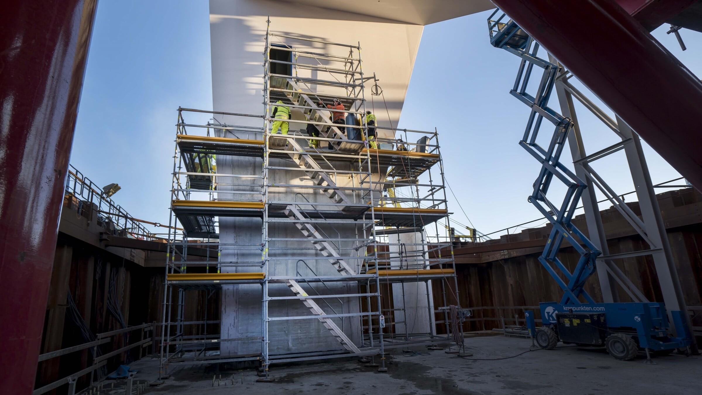 Byggnadsarbetare på byggnadsställningar nära en stor betongstruktur med en blå lift i närheten.