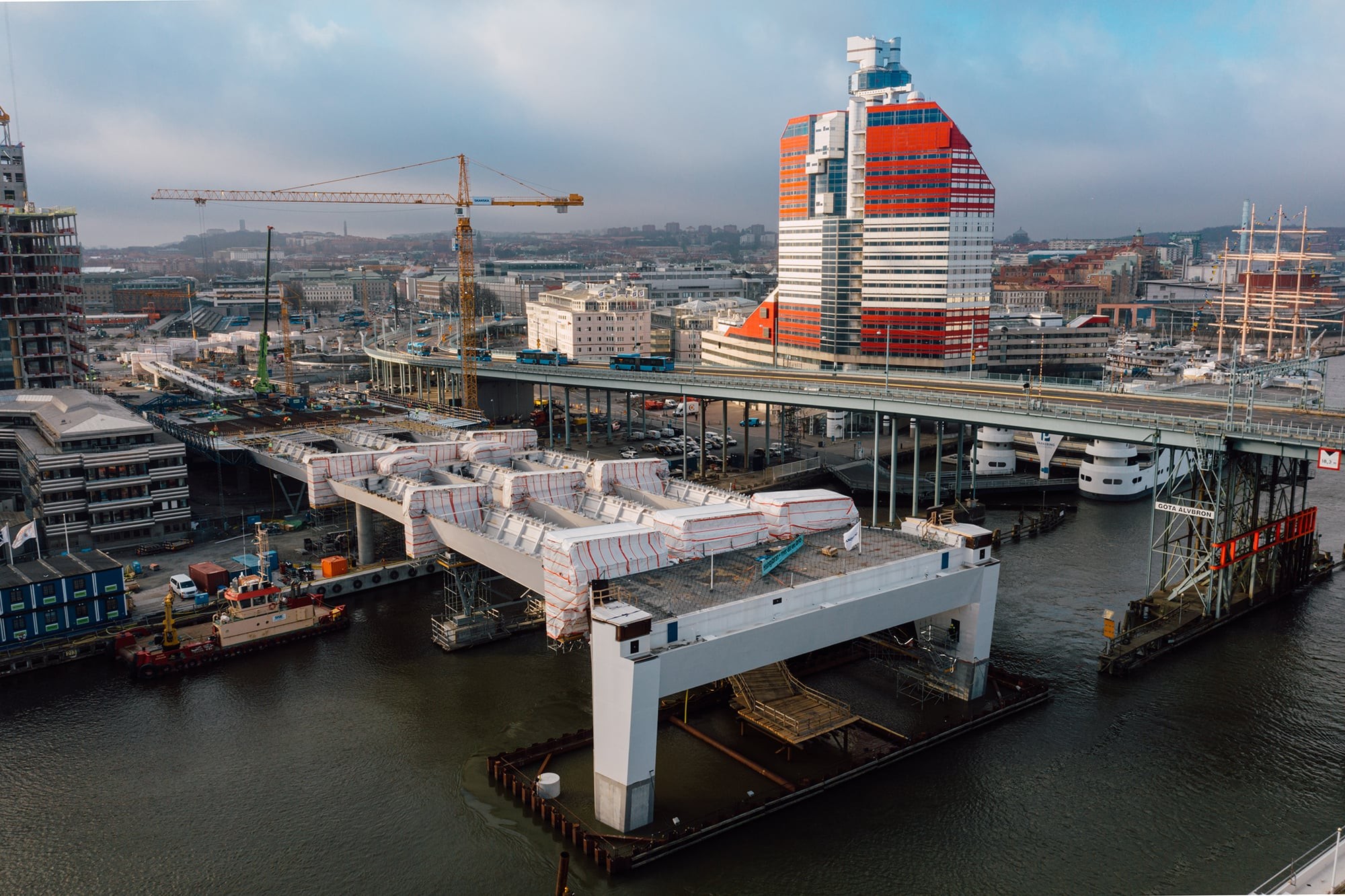 Brobygge över en flod med kranar, intill en färgglad modern byggnad i ett stadsområde.