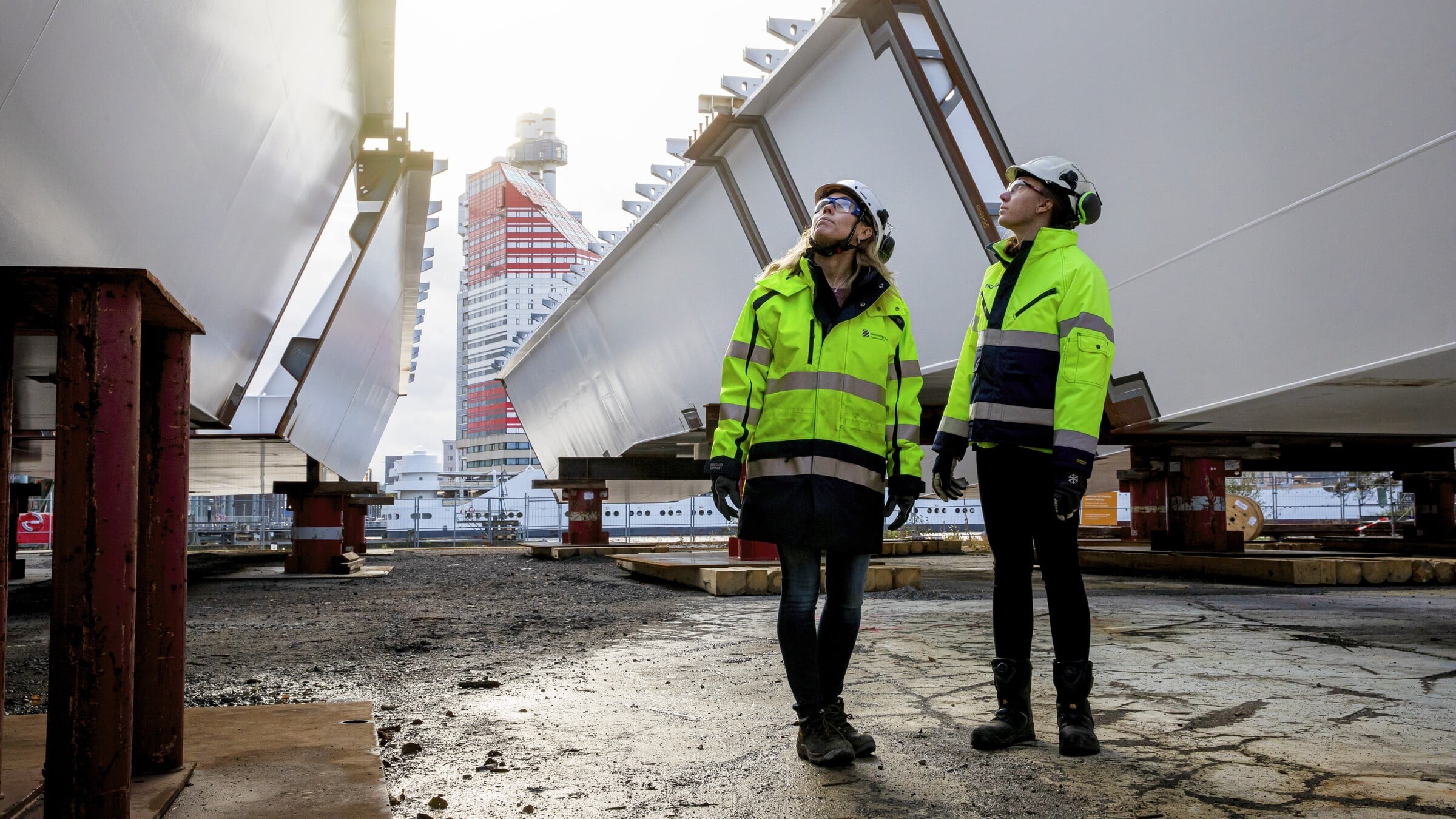 Två arbetare i skyddsutrustning inspekterar stora industriella strukturer i en utomhusmiljö.
