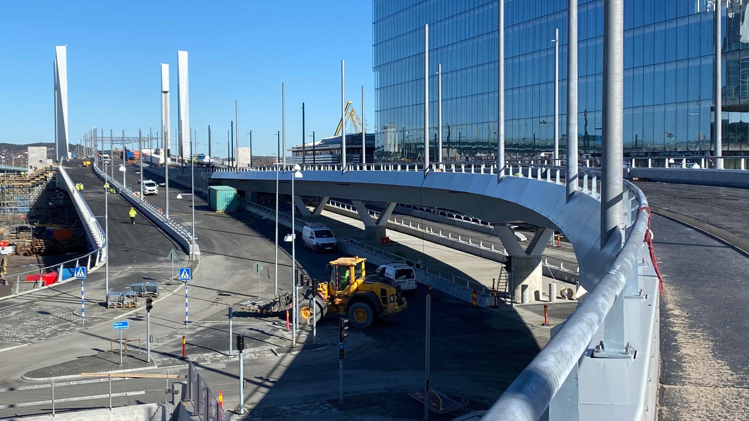 Modern brokonstruktion med fordon och maskineri nära en glasbyggnad i stadsområde.