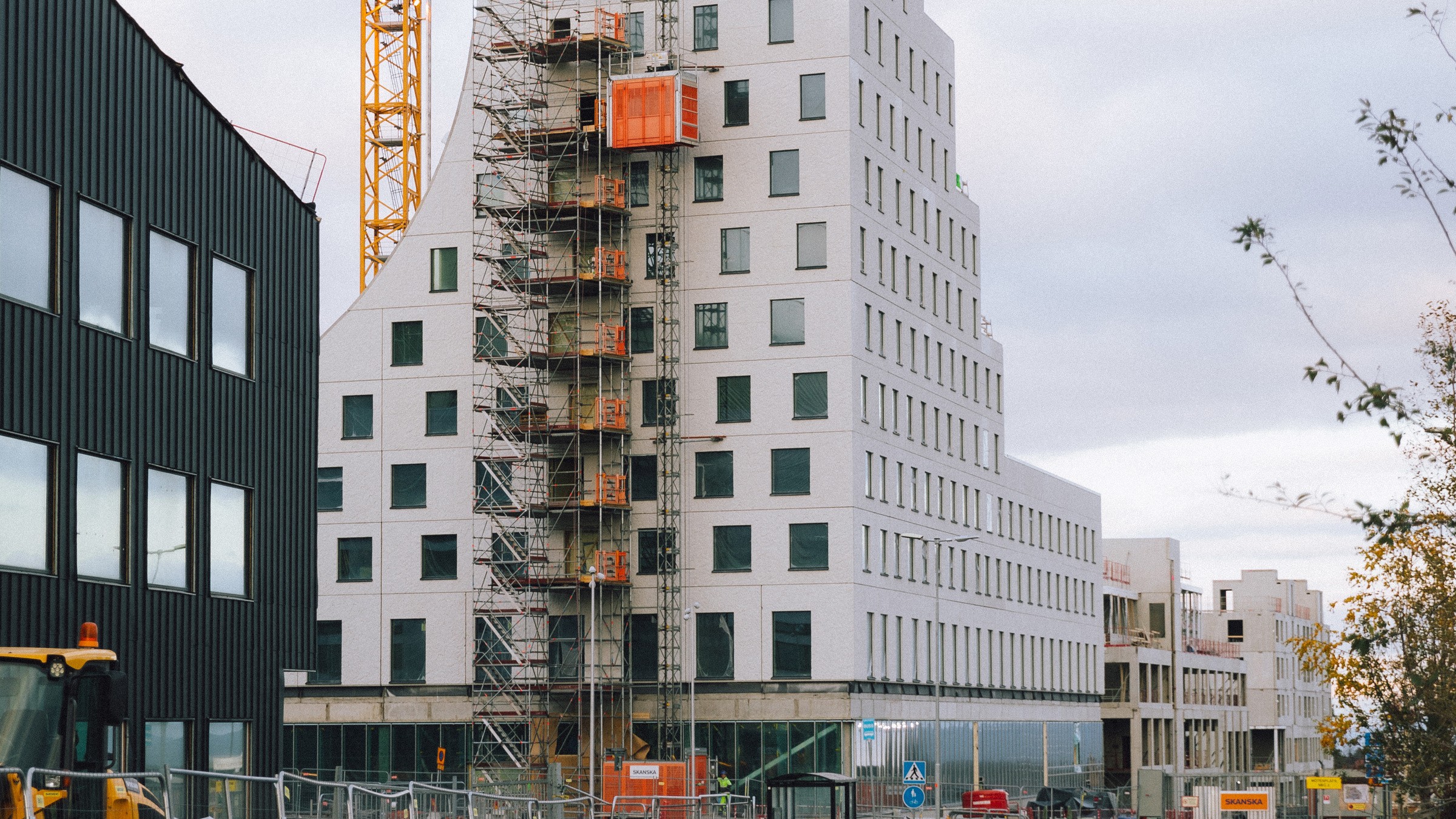 Modern byggnad under konstruktion med byggnadsställningar och kran, omgiven av stadslandskap.