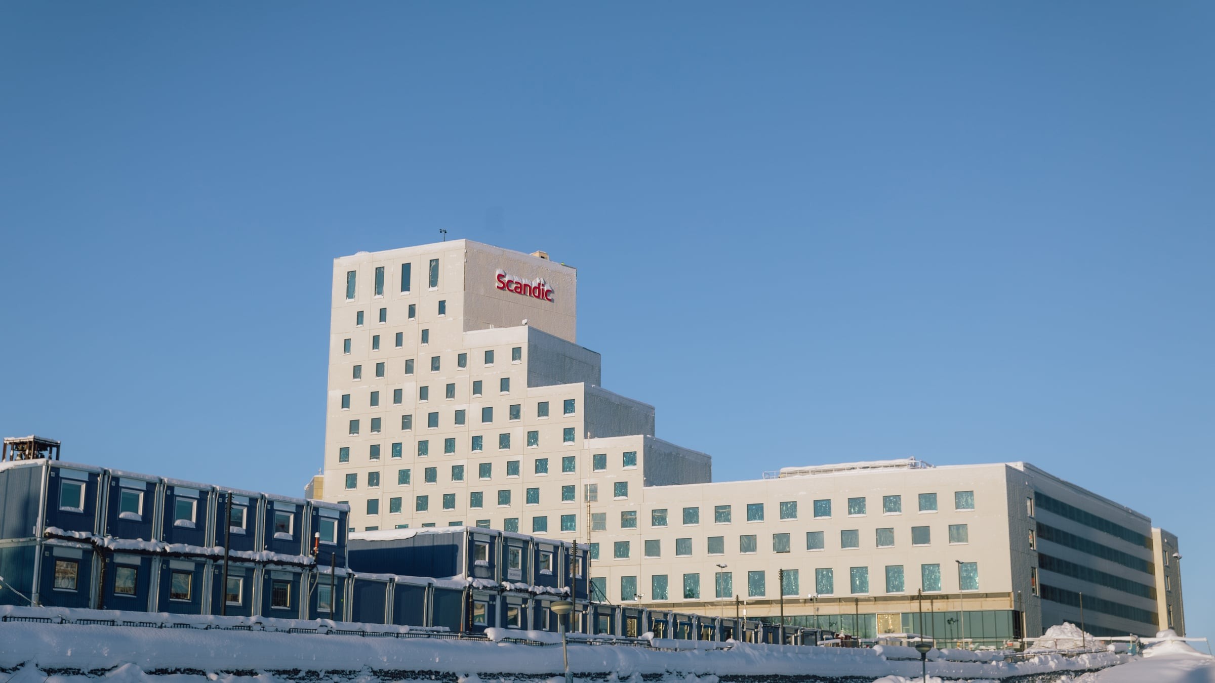 Modern Scandic hotellbyggnad med snöiga omgivningar under en klarblå himmel.