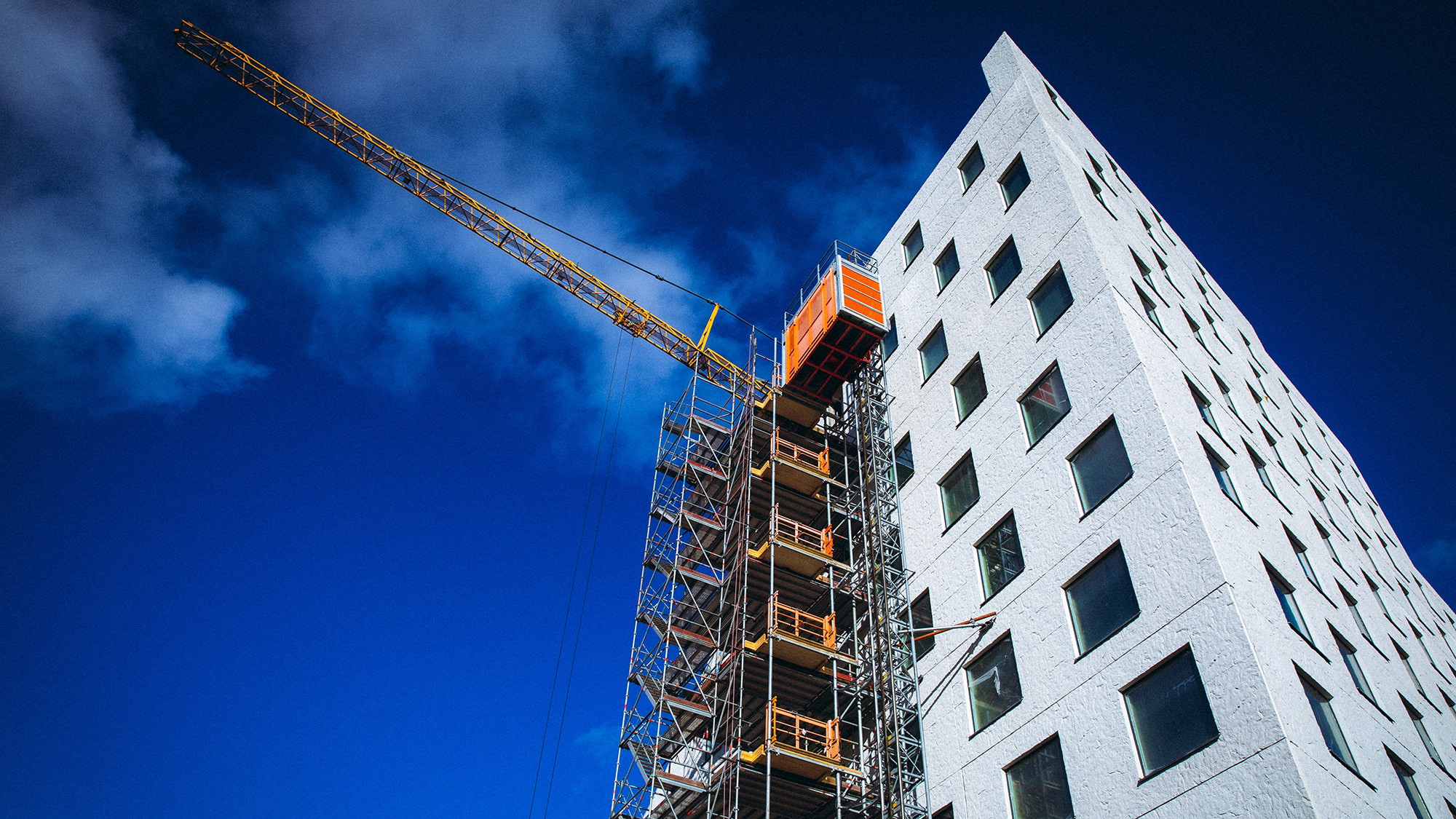 En modern vit byggnad under konstruktion med byggnadsställningar och en kran mot en klarblå himmel.
