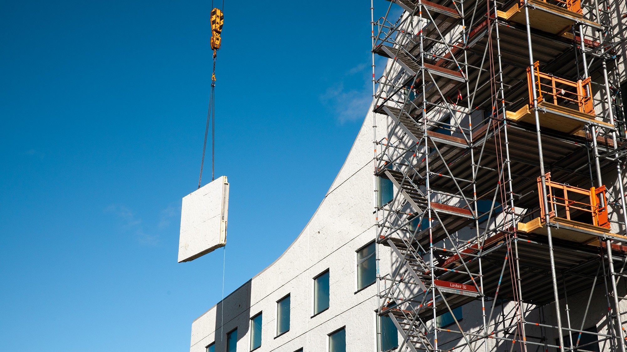 Byggarbetsplats med byggnadsställningar och en kran som lyfter en betongpanel mot en blå himmel.