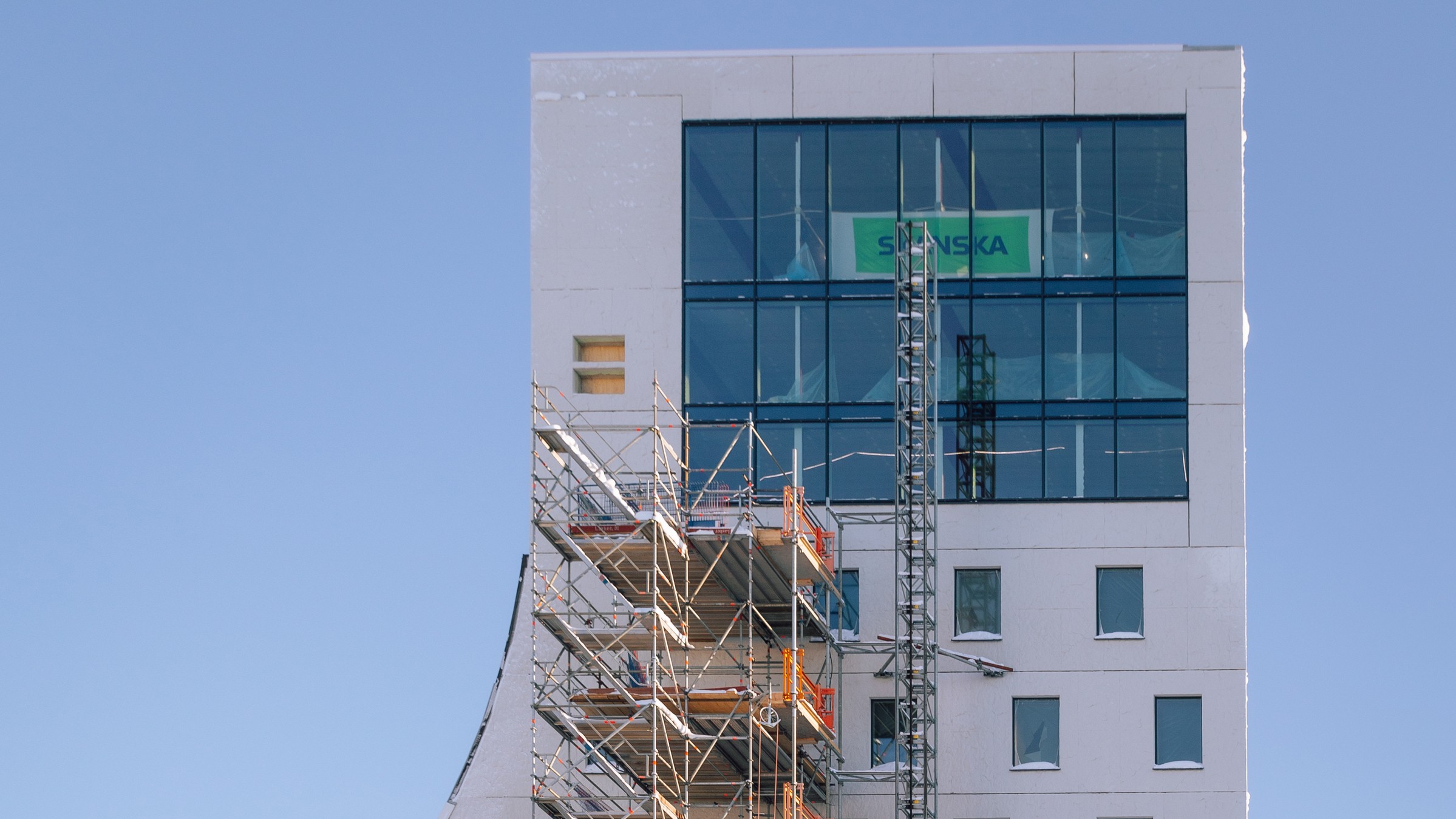 Byggnad under konstruktion med byggnadsställningar och stora glasfönster mot en klarblå himmel.