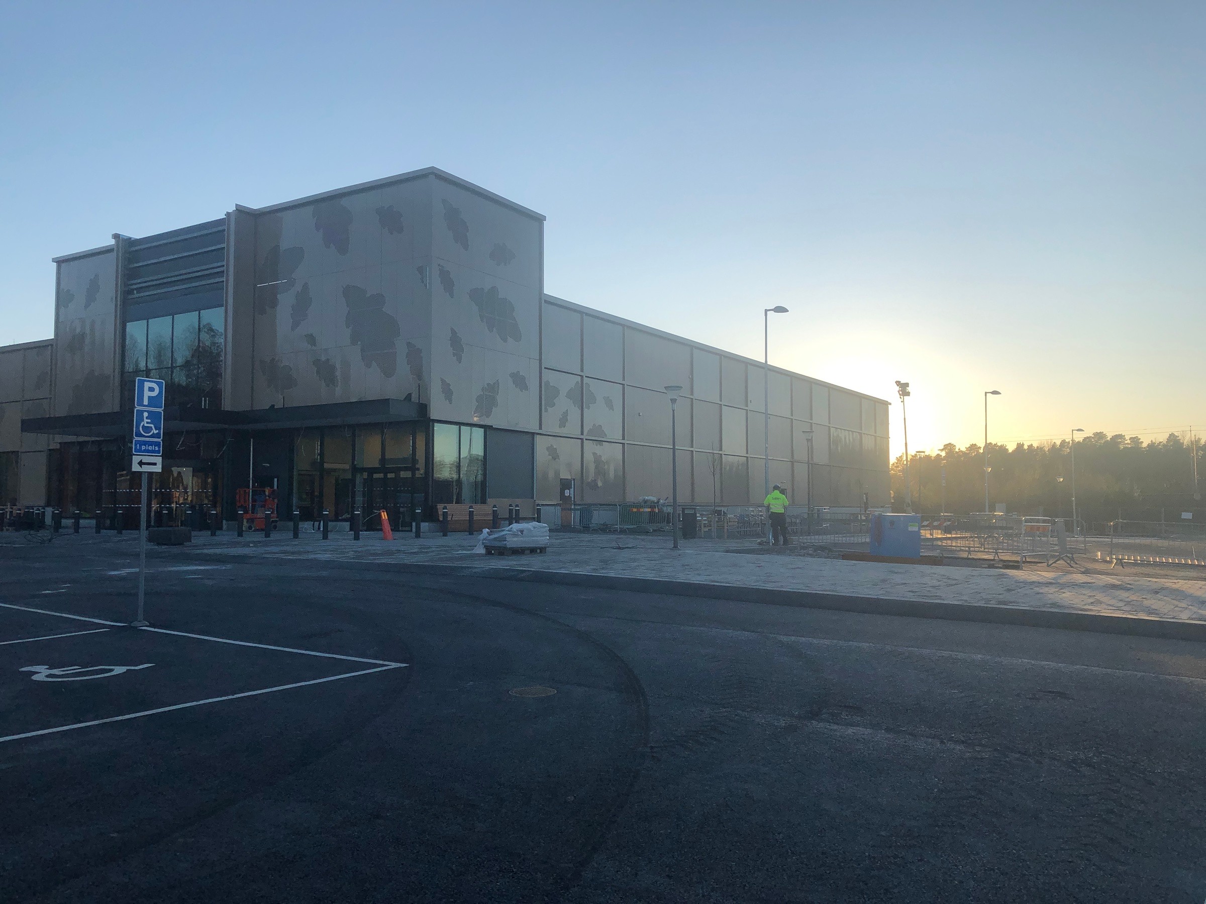Skanska har utvecklat en ny ICA Maxi Stormarknad i Bålsta. Modern kommersiell byggnad med glasfasad på en parkeringsplats vid soluppgången, med tillgänglighetsskyltar.