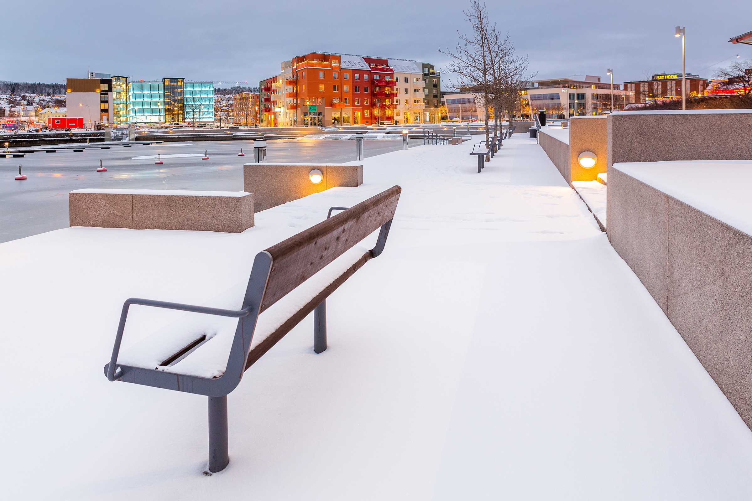 Snötäckt stadspark med moderna byggnader i bakgrunden, med en bänk och en gångväg.