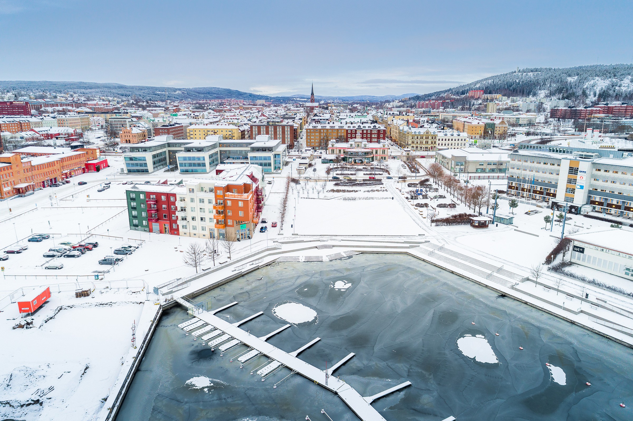 Flygfoto av en snötäckt stadssilhuett med färgglada byggnader och frusen vattenfront, mot en bakgrund av kullar.