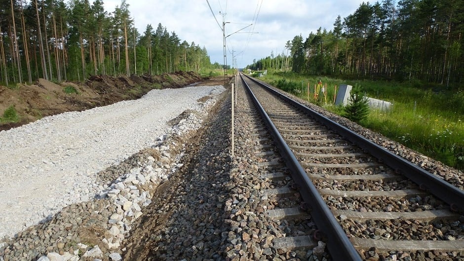 Järnvägsspår bredvid en grusstig i ett frodigt skogsområde under en delvis molnig himmel.